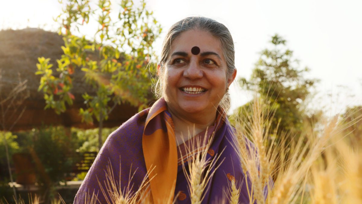 fotografi av personen vandana shiva som står i en åker med fjell og trær i bakgrunnen