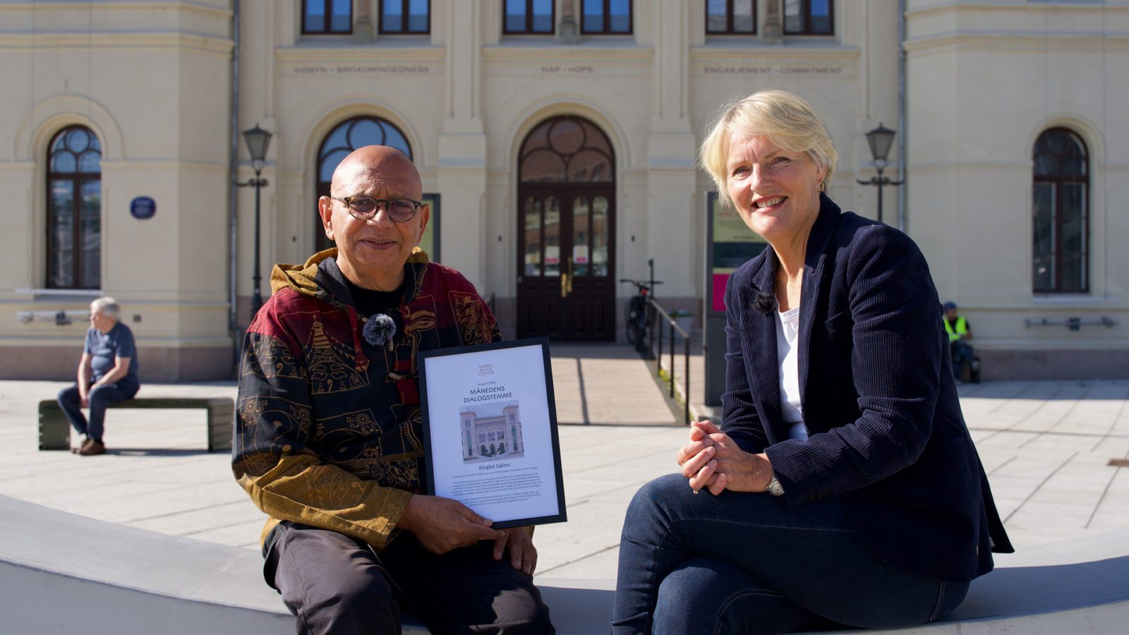 Khalid Salimi sitter med et diplom i hånden. Ved siden av ham sitter Kjersti Fløgstad. Begge sitter med ryggen til Nobels fredssenter