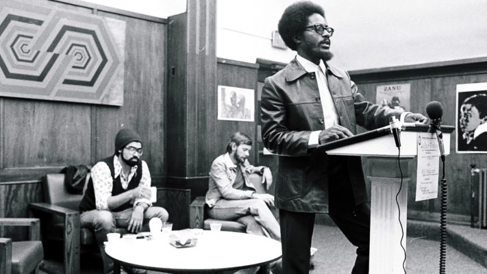 Walter Rodney står ved en talerstol. To menn sitter i bakgrunnen